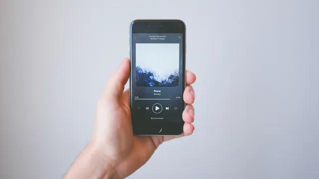Mão a segurar um iPhone com o leitor do Spotify aberto, a mostrar a capa de um álbum e os botões de reprodução, sobre fundo cinzento