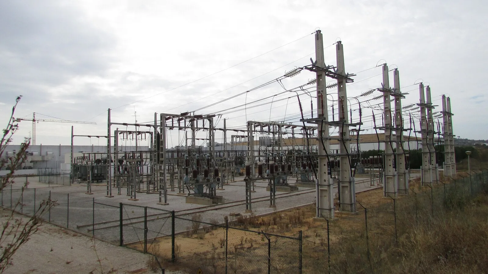 Subestação de distribuição de eletricidade ao ar livre, com postes de betão, isoladores e cabos, vedada por uma rede, sob céu nublado