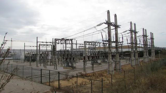 Subestação de distribuição de eletricidade ao ar livre, com postes de betão, isoladores e cabos, vedada por uma rede, sob céu nublado