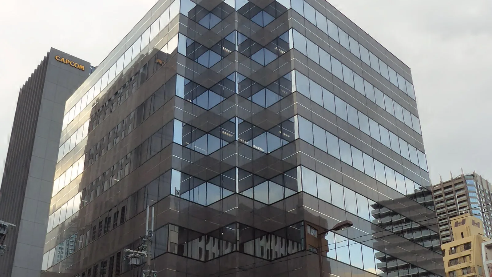 Torre cinzenta com o letreiro CAPCOM no topo, ao lado de um edifício de escritórios com fachada de vidro espelhado, numa rua de Osaka