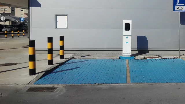 Posto de carregamento branco da rede MOBI.E encostado à parede de um supermercado, com dois lugares de estacionamento pintados de azul e pilaretes amarelos e pretos