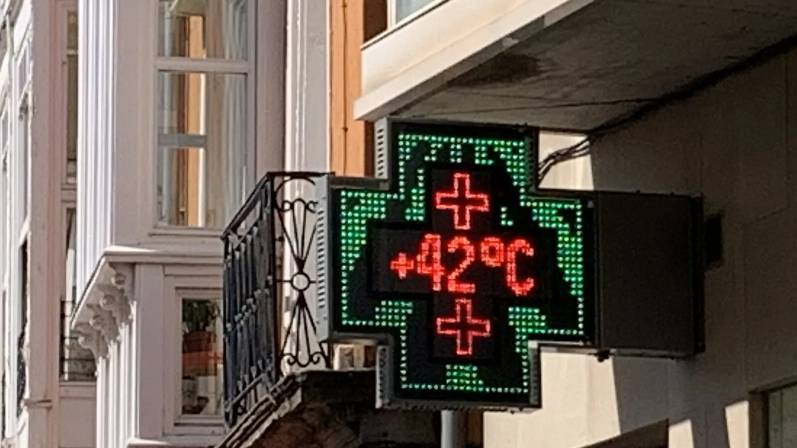 Cruz verde luminosa de uma farmácia presa a uma fachada, a mostrar +42 °C em letras vermelhas, numa rua com varandas envidraçadas