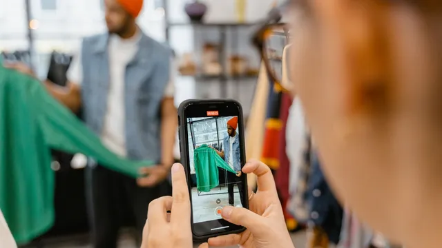 Mão a segurar um telemóvel apontado a um homem que mostra uma camisa verde numa loja de roupa, com a imagem da camisa no ecrã