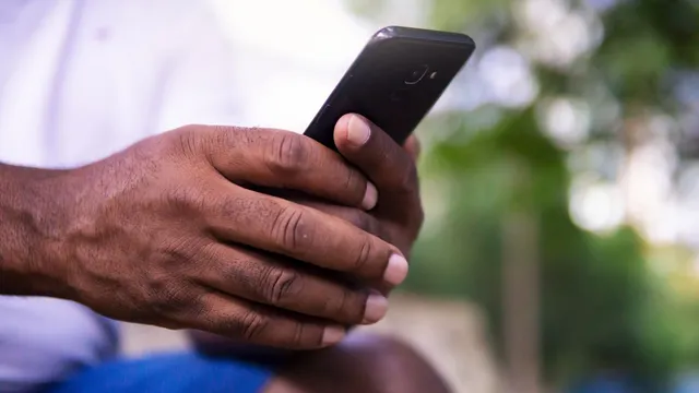 Mãos de um homem a segurar um telemóvel preto ao ar livre, com árvores desfocadas ao fundo
