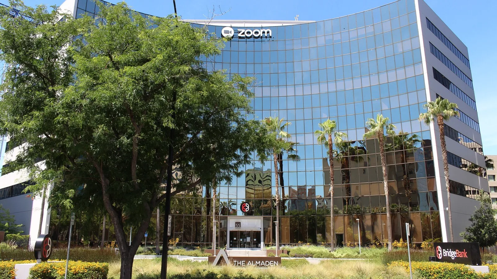 Edifício envidraçado da sede da Zoom em San José, na Califórnia, com o logótipo da empresa no topo, árvores e palmeiras à frente