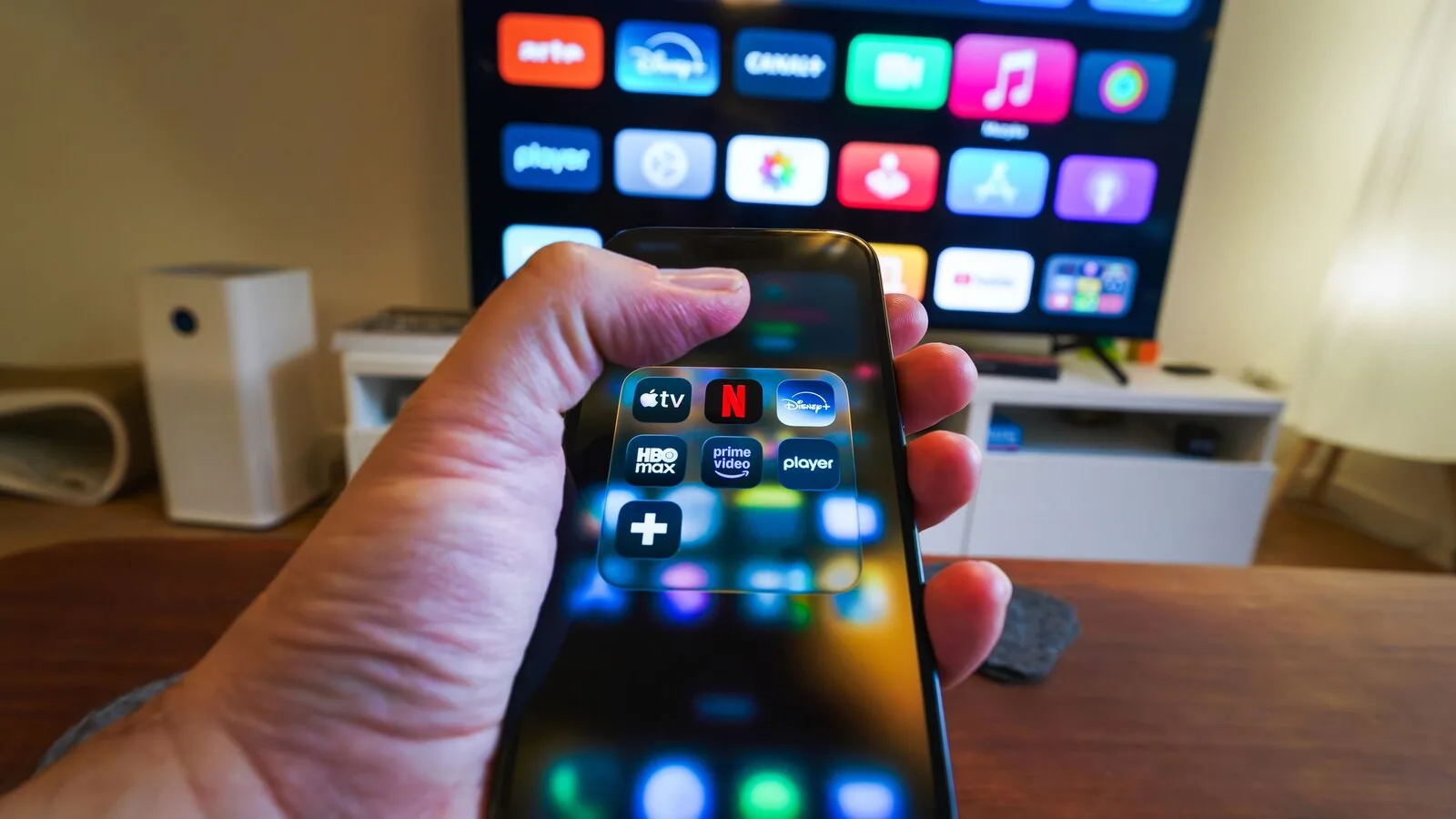 Mão a segurar um telemóvel com ícones de apps de streaming, em frente a uma televisão com uma grelha de aplicações no ecrã inicial