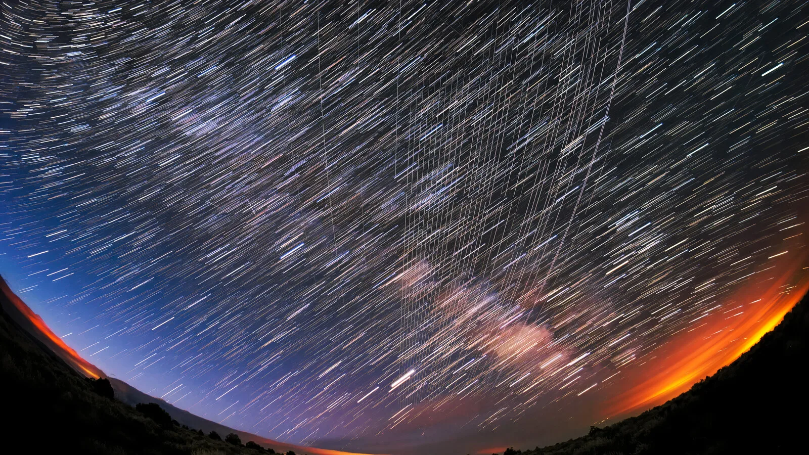 Céu noturno numa fotografia de longa exposição, com rastos de estrelas em arco e linhas paralelas deixadas por satélites Starlink