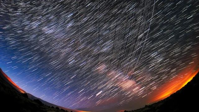 Céu noturno numa fotografia de longa exposição, com rastos de estrelas em arco e linhas paralelas deixadas por satélites Starlink
