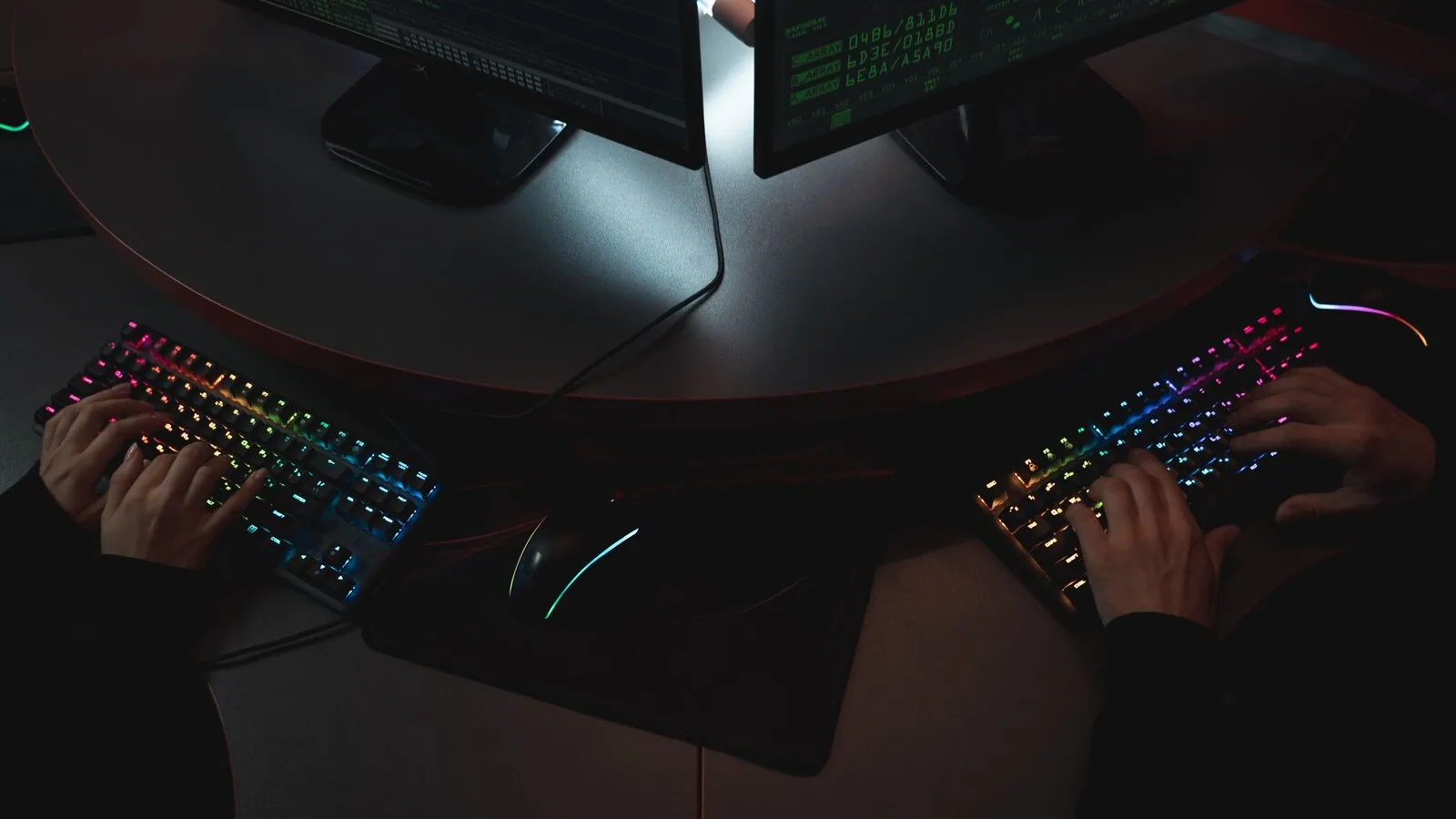 Duas pessoas a escrever em teclados iluminados, numa sala escura com vários monitores