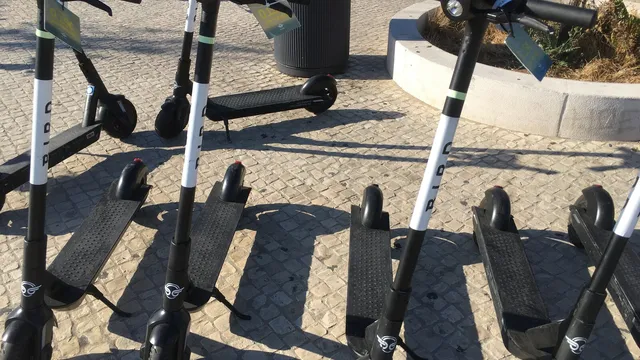 Várias trotinetes elétricas partilhadas, pretas e brancas, estacionadas na calçada portuguesa em Lisboa, ao sol