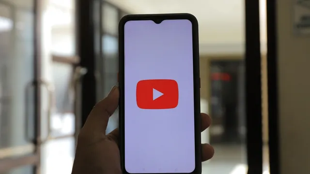 Mão a segurar um telemóvel com o logótipo do YouTube no ecrã, junto a uma porta de vidro