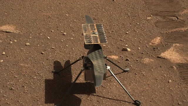 O helicóptero Ingenuity pousado no solo avermelhado e pedregoso de Marte, com o painel solar no topo, as duas pás alinhadas e as quatro pernas finas a projetar sombra