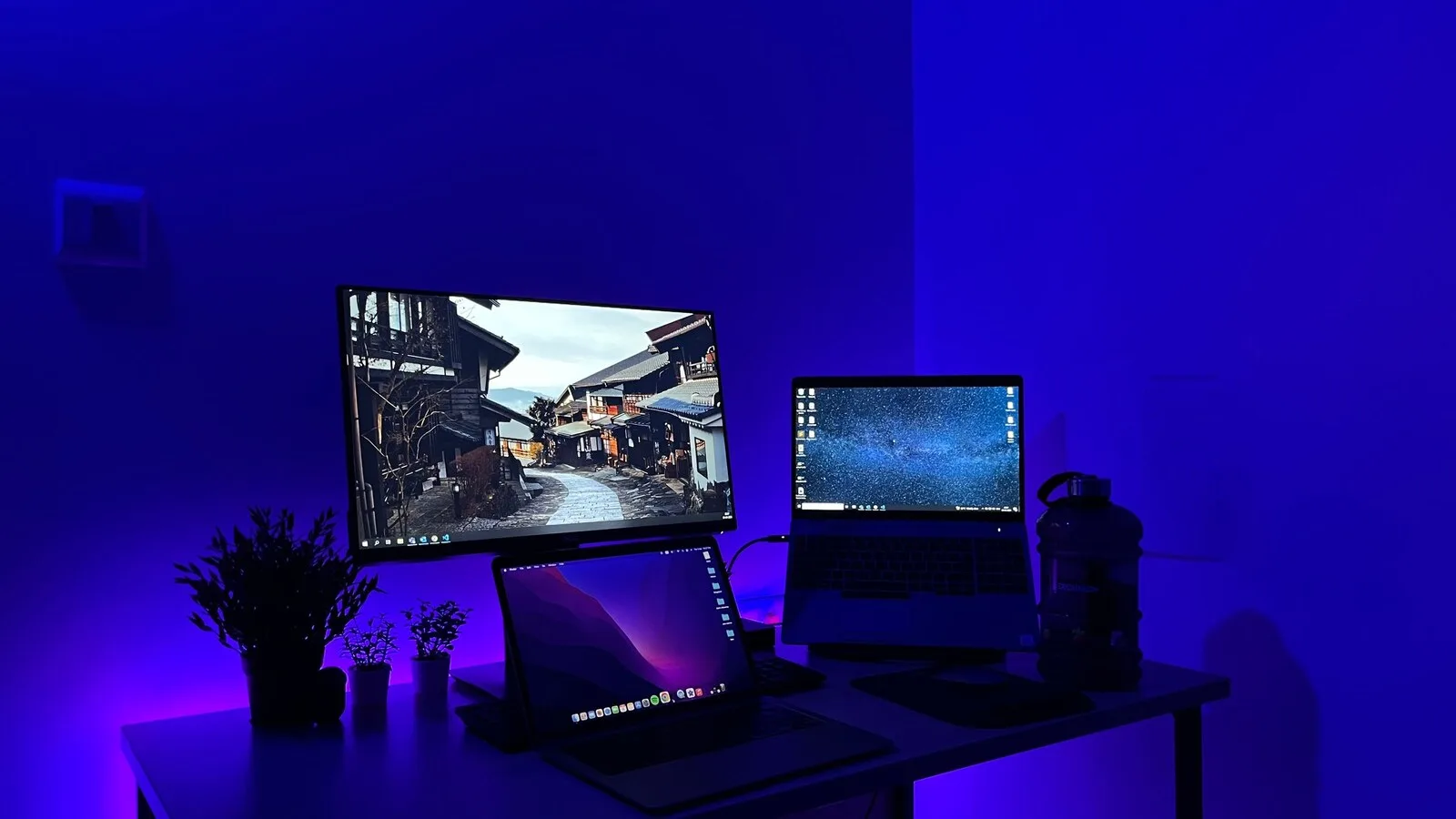 Secretária iluminada a azul com um monitor e dois portáteis ligados, um deles com o ambiente de trabalho do Windows
