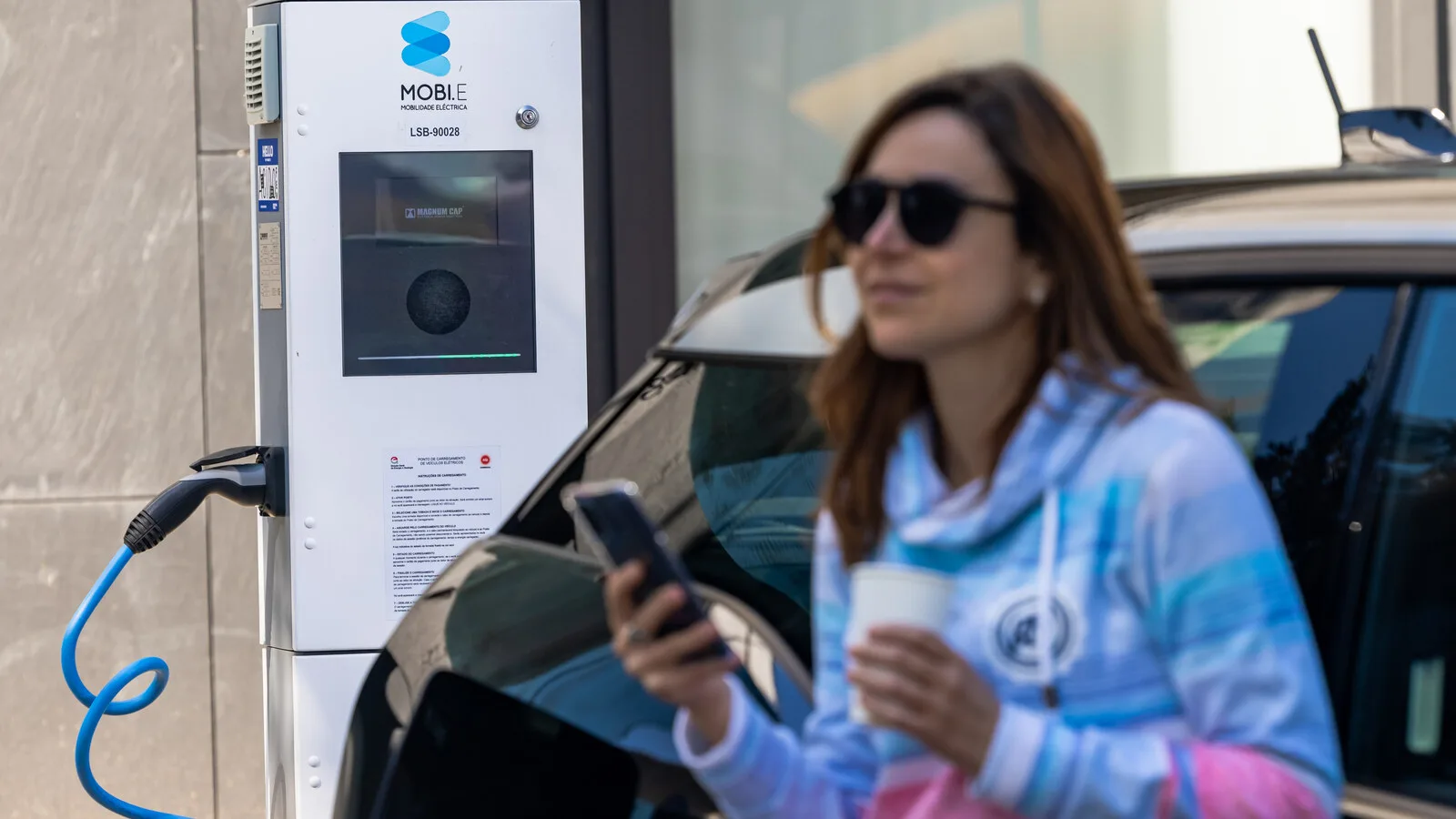 Mulher com um telemóvel e um copo de café encostada a um carro ligado a um posto de carregamento da rede MOBI.E