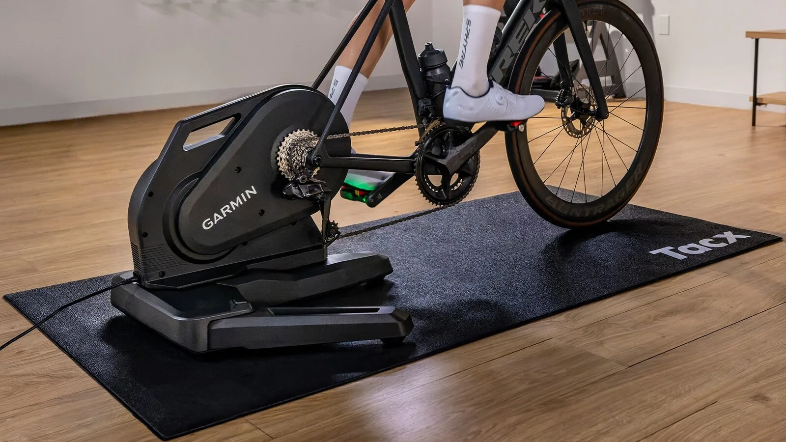 Ciclista a pedalar numa bicicleta de estrada montada no rolo de treino Garmin Tacx Nēra, preto, pousado sobre um tapete numa sala com chão de madeira