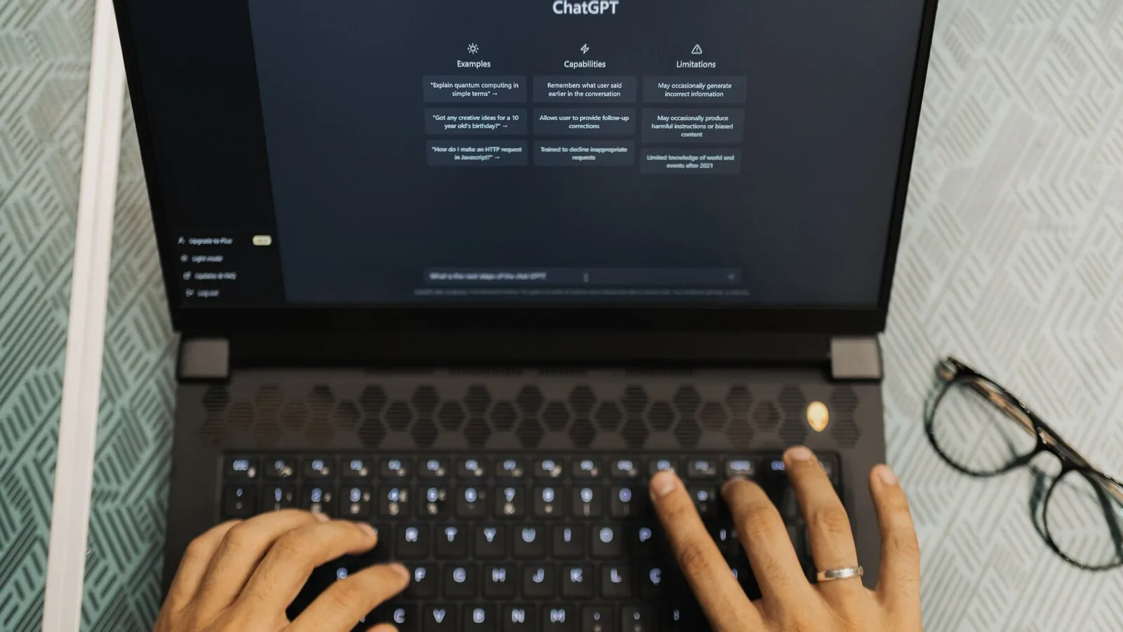Mãos a escrever no teclado de um portátil com a página inicial do ChatGPT aberta no ecrã e uns óculos ao lado