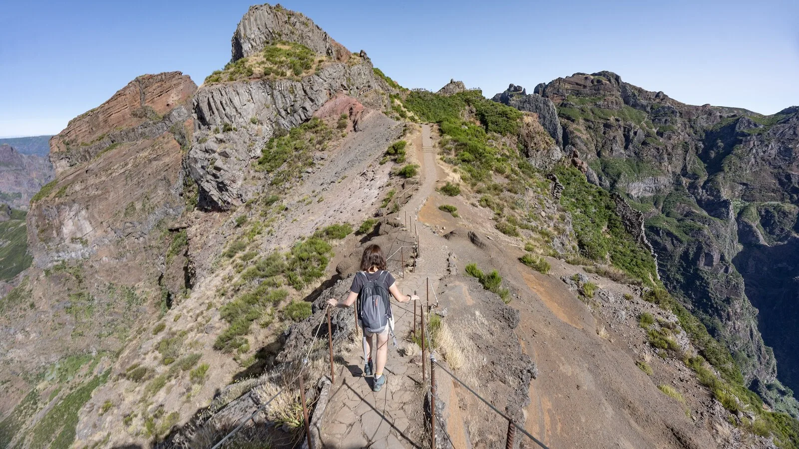 Caminhante de mochila num trilho de montanha estreito com corrimão, entre picos rochosos, na Madeira
