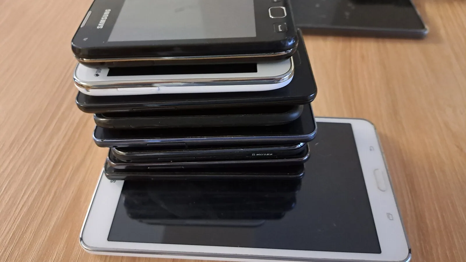 Pilha de oito telemóveis Samsung antigos, pretos e um branco, pousada sobre um tablet branco numa mesa de madeira