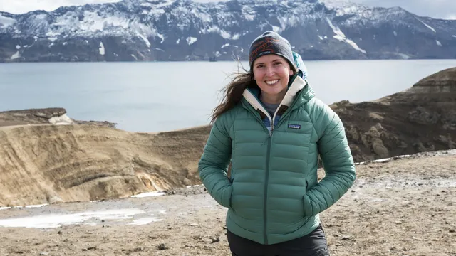 Mulher sorridente com casaco verde e gorro, de pé num terreno vulcânico, com um lago e montanhas com neve ao fundo