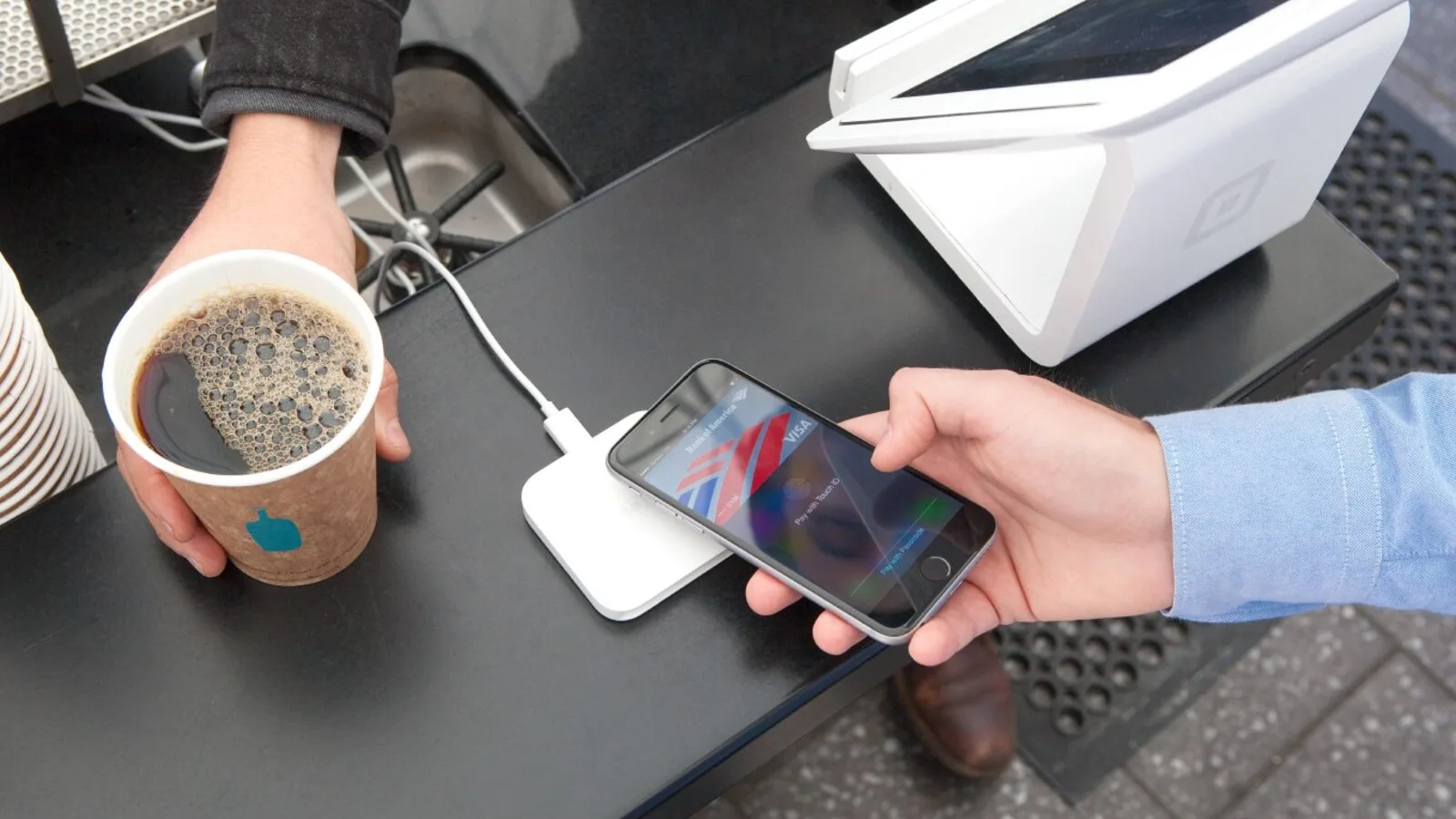 Homem com um copo de café na mão encosta um iPhone a um leitor de pagamentos branco, num balcão preto