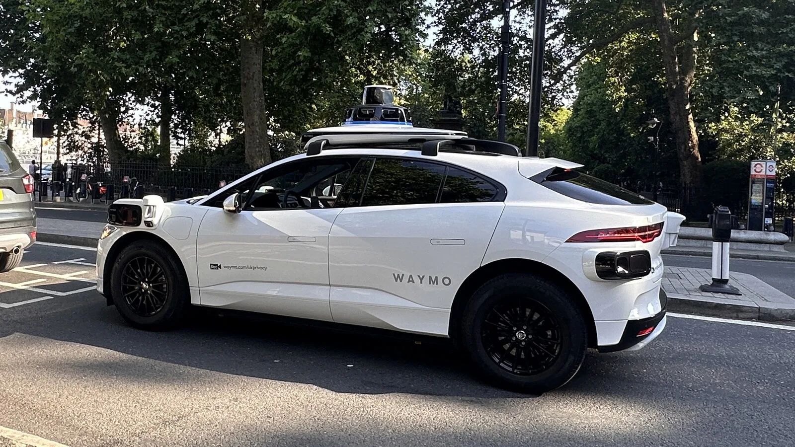 Carro elétrico branco da Waymo com sensores e câmaras no tejadilho, parado numa rua arborizada de Londres