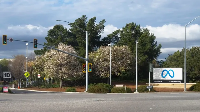 Placa com o logótipo da Meta e a morada 1 Hacker Way à entrada da sede da empresa em Menlo Park, junto a um cruzamento com semáforos