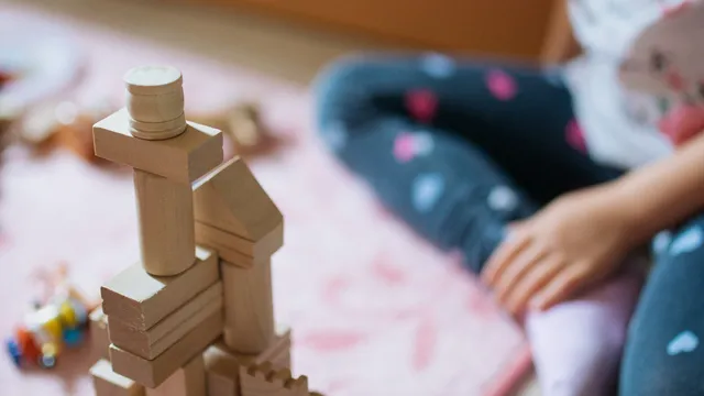Torre de blocos de madeira em primeiro plano, com uma criança sentada num tapete cor-de-rosa desfocada ao fundo