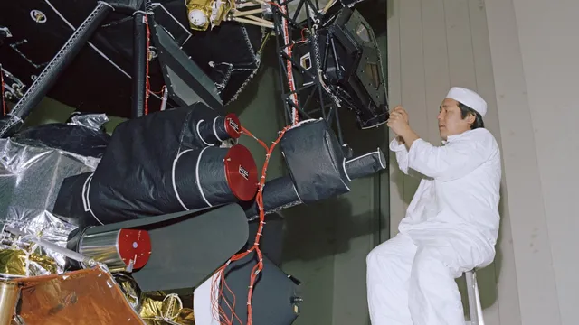Técnico de fato branco, sentado num escadote, a ajustar instrumentos científicos de uma sonda Voyager, com cilindros pretos de tampa vermelha e isolamento dourado
