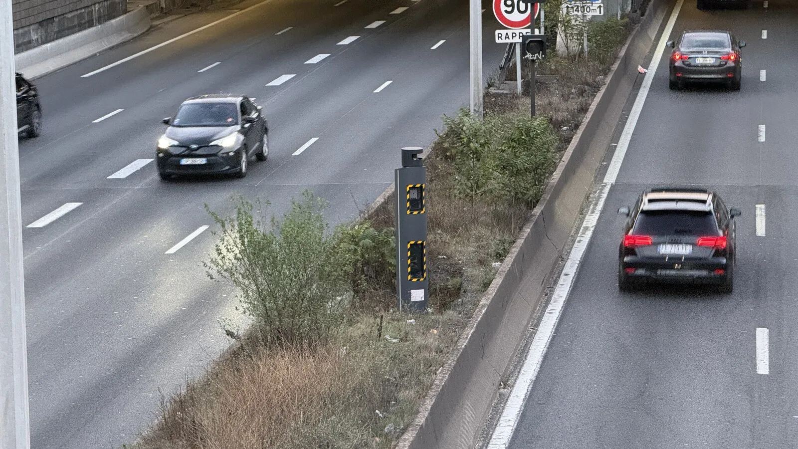 Radar de velocidade cinzento com faixas amarelas no separador central de uma autoestrada, com carros a passar nos dois sentidos e um sinal de limite de 90 ao fundo