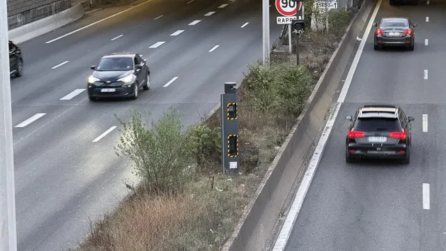 Radar de velocidade cinzento com faixas amarelas no separador central de uma autoestrada, com carros a passar nos dois sentidos e um sinal de limite de 90 ao fundo
