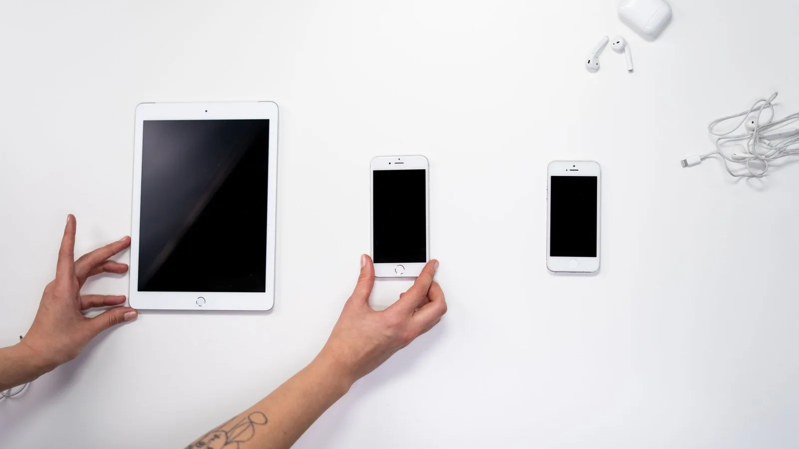 Mãos a arrumar um iPad e dois iPhone numa mesa branca, com uns AirPods e o estojo no canto