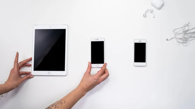 Mãos a arrumar um iPad e dois iPhone numa mesa branca, com uns AirPods e o estojo no canto