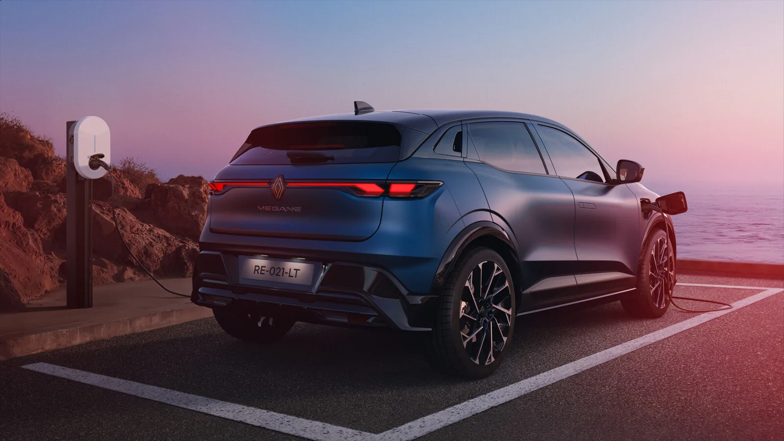 Renault Mégane E-Tech azul-escuro estacionado ao entardecer, ligado por cabo a um posto de carregamento, com falésias e o mar ao fundo