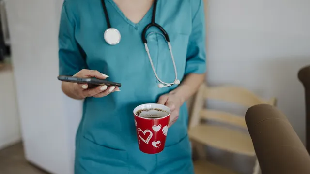 Pessoa de farda hospitalar com estetoscópio a consultar o telemóvel com uma caneca de café vermelha na outra mão