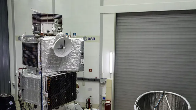 Sonda espacial coberta de isolamento prateado, com painéis solares escuros e uma antena parabólica branca, montada num pavilhão de testes com o logótipo da ESA na parede
