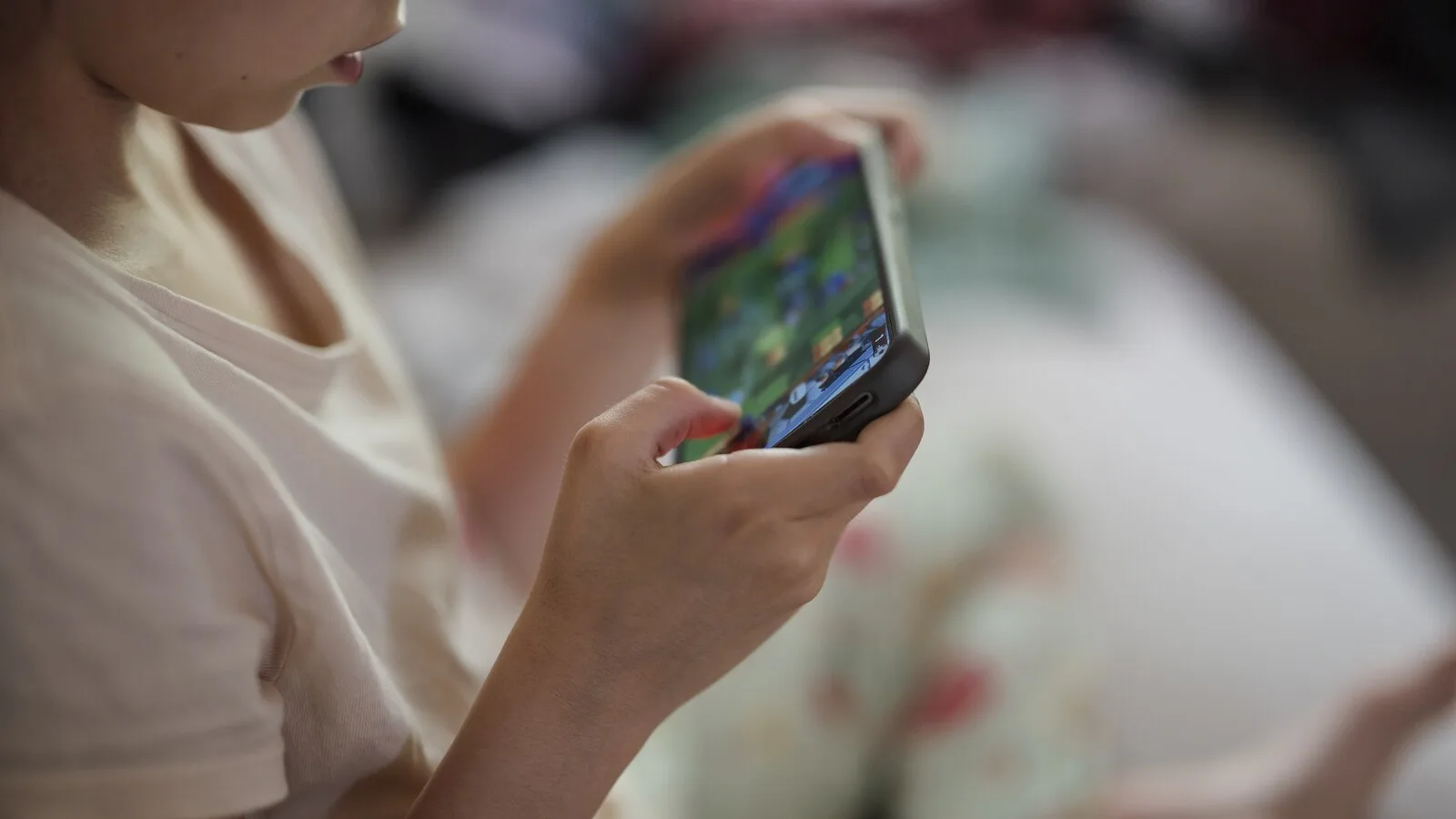 Criança de t-shirt clara a jogar num telemóvel que segura com as duas mãos, sentada numa cama