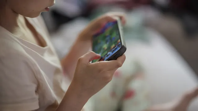 Criança de t-shirt clara a jogar num telemóvel que segura com as duas mãos, sentada numa cama