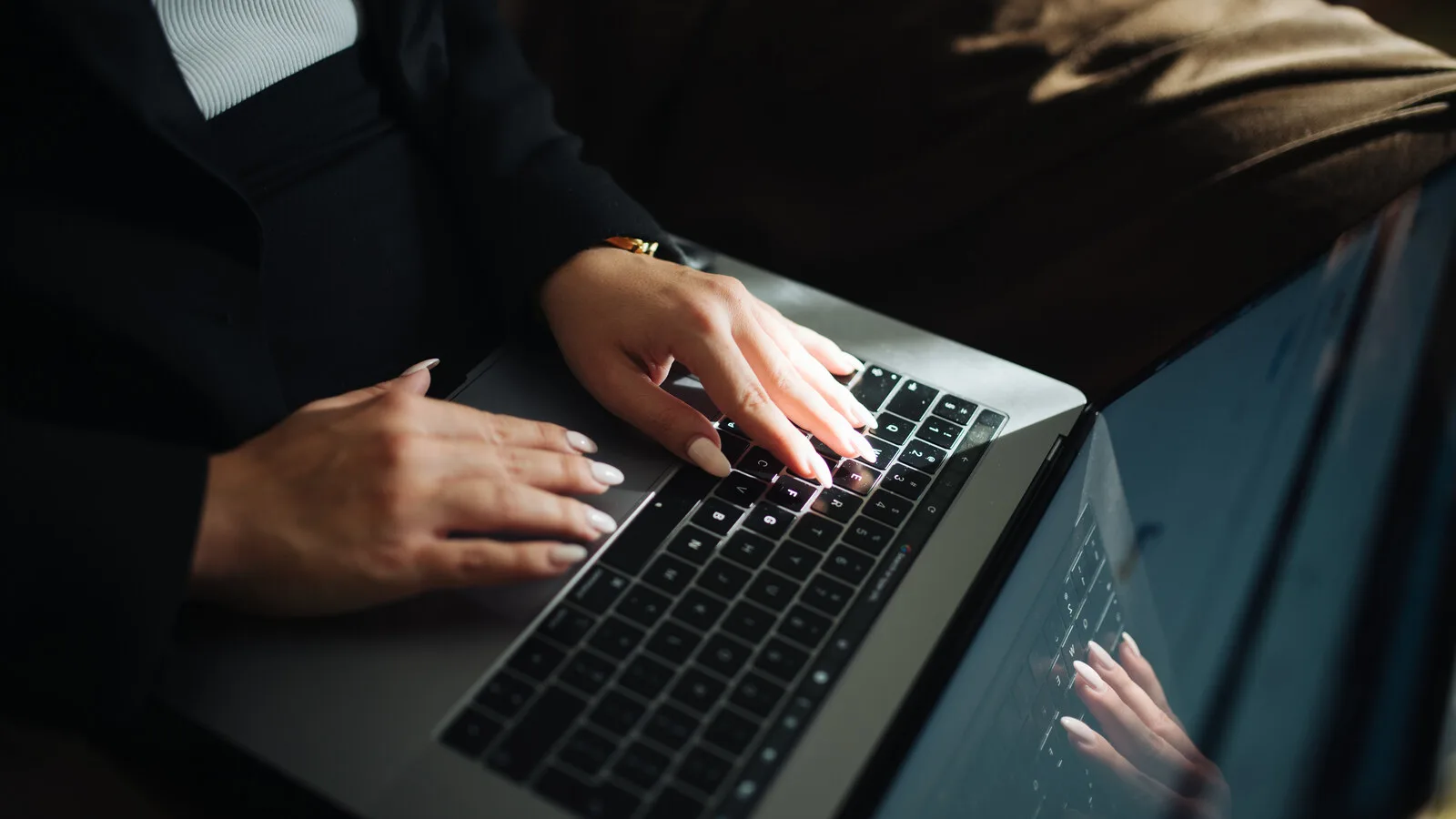 Mãos de uma mulher de unhas claras a escrever no teclado iluminado de um portátil, com o reflexo no ecrã