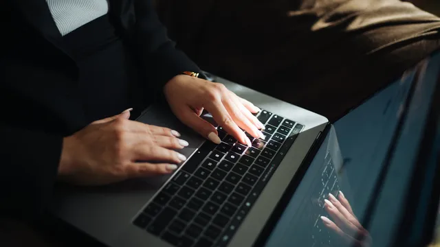 Mãos de uma mulher de unhas claras a escrever no teclado iluminado de um portátil, com o reflexo no ecrã