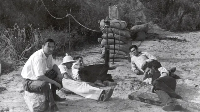Fotografia a preto e branco de cinco jovens sentados e deitados na areia junto a um motor de foguetão montado entre sacos de areia