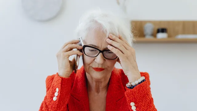 Mulher de cabelo branco e óculos, com casaco vermelho, ao telemóvel com a mão na testa e ar preocupado