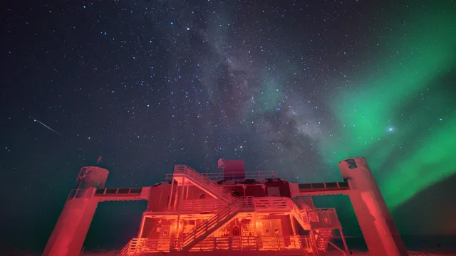 Edifício do laboratório IceCube iluminado a vermelho na noite polar, com a Via Láctea e uma aurora verde no céu