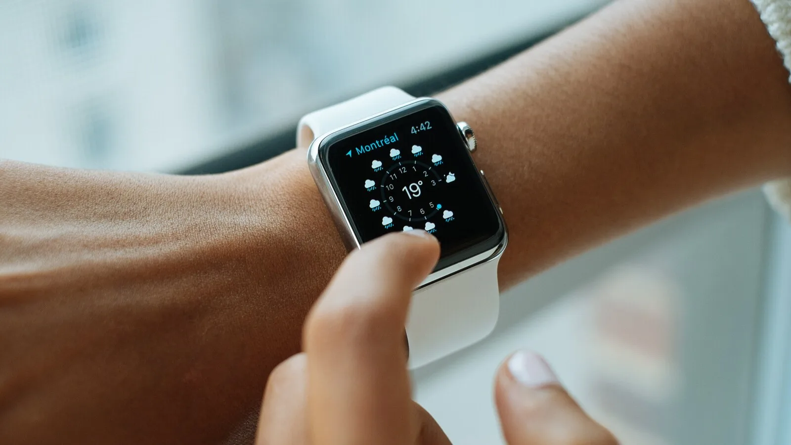 Pessoa toca com o dedo no ecrã de um Apple Watch com bracelete branca que mostra a previsão do tempo
