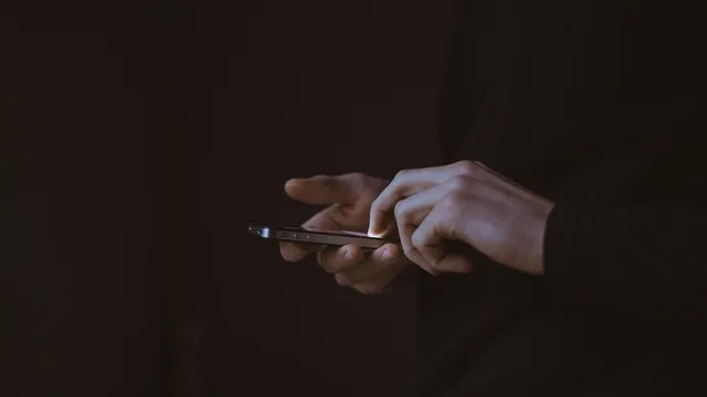 Mãos a escrever num telemóvel, com o ecrã a iluminar os dedos num fundo escuro