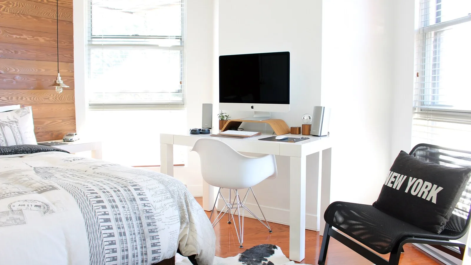 Quarto luminoso com cama de edredão estampado, secretária branca com um computador e uma cadeira preta com uma almofada
