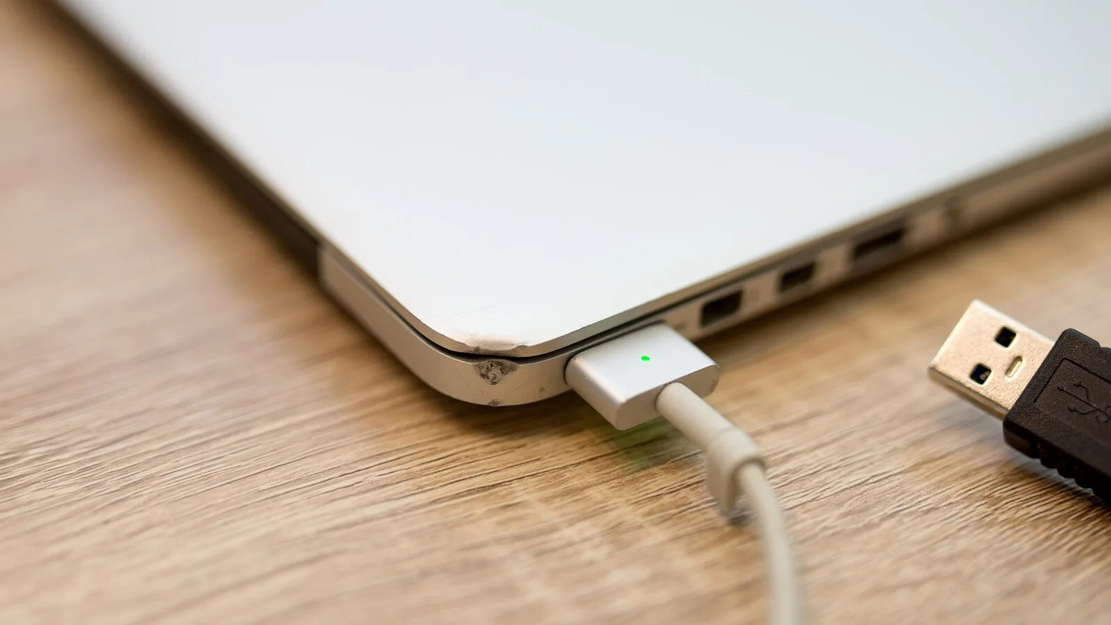 Canto de um portátil prateado ligado a um carregador magnético com a luz verde acesa, sobre uma mesa de madeira, com uma ficha USB ao lado
