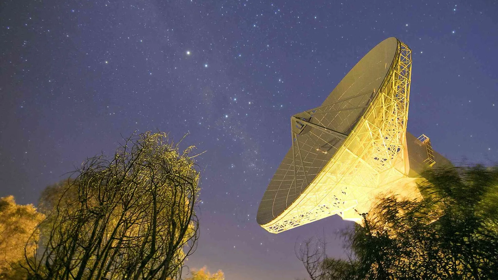 Grande antena parabólica de uma estação terrestre de comunicações com satélites, iluminada à noite sob um céu estrelado, com árvores em primeiro plano