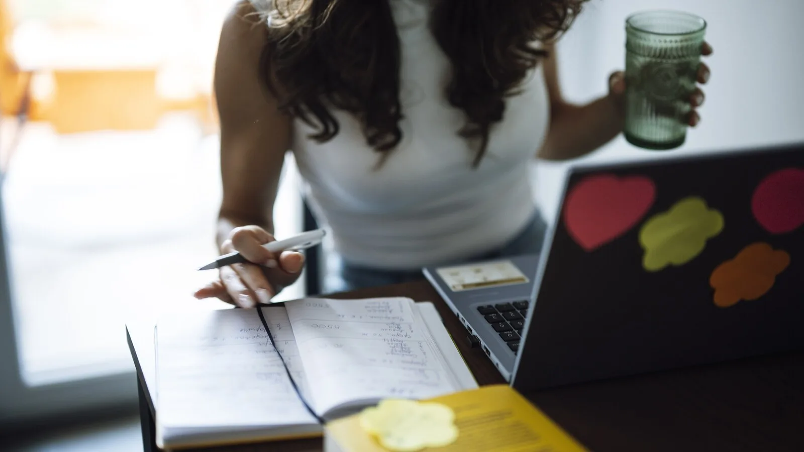 Mulher a estudar numa secretária com um caderno de apontamentos aberto, uma caneta na mão, um portátil com autocolantes e um copo de água