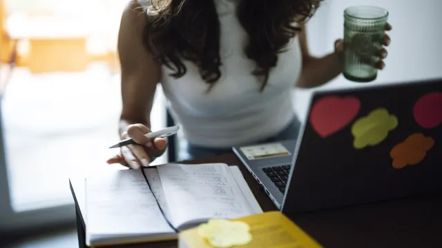Mulher a estudar numa secretária com um caderno de apontamentos aberto, uma caneta na mão, um portátil com autocolantes e um copo de água