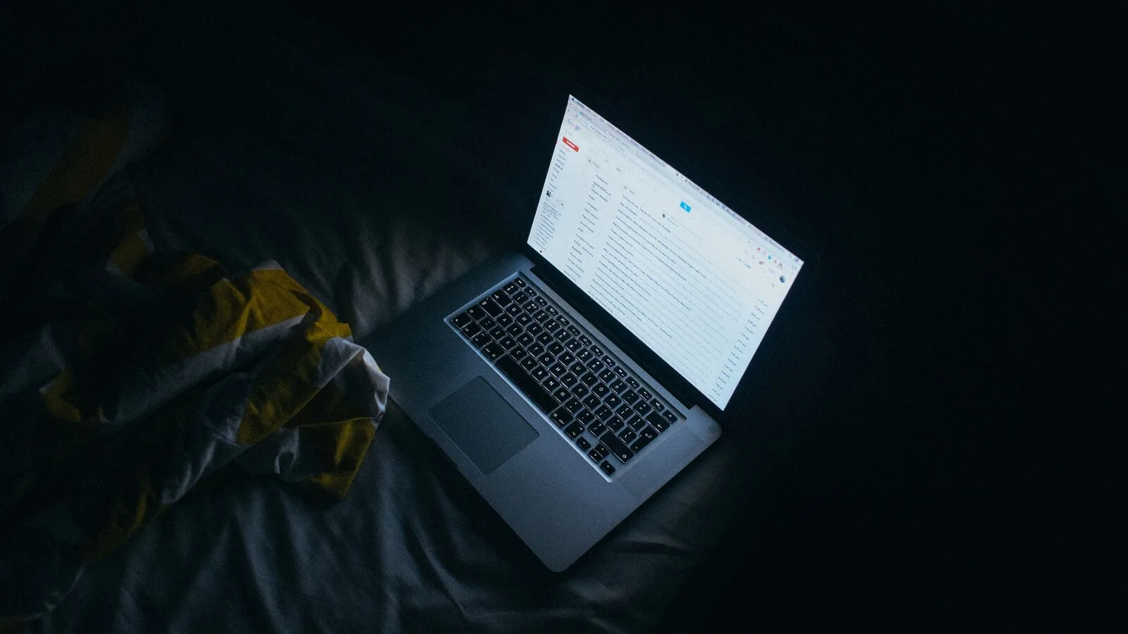 Portátil aberto em cima de uma cama, num quarto às escuras, com o ecrã muito branco a mostrar uma caixa de email